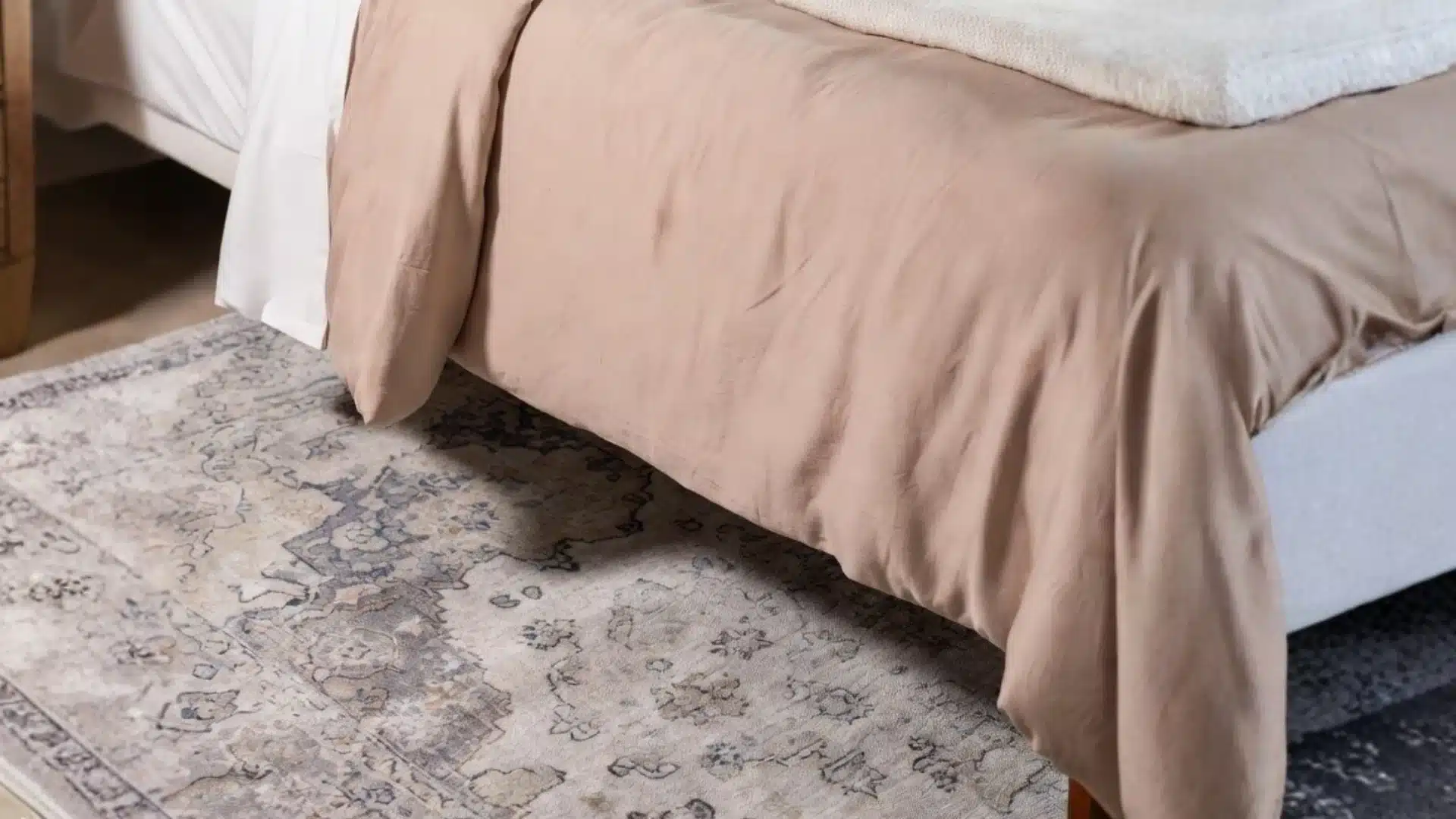 A close up of a bed with a tan duvet cover resting above a light colored area rug