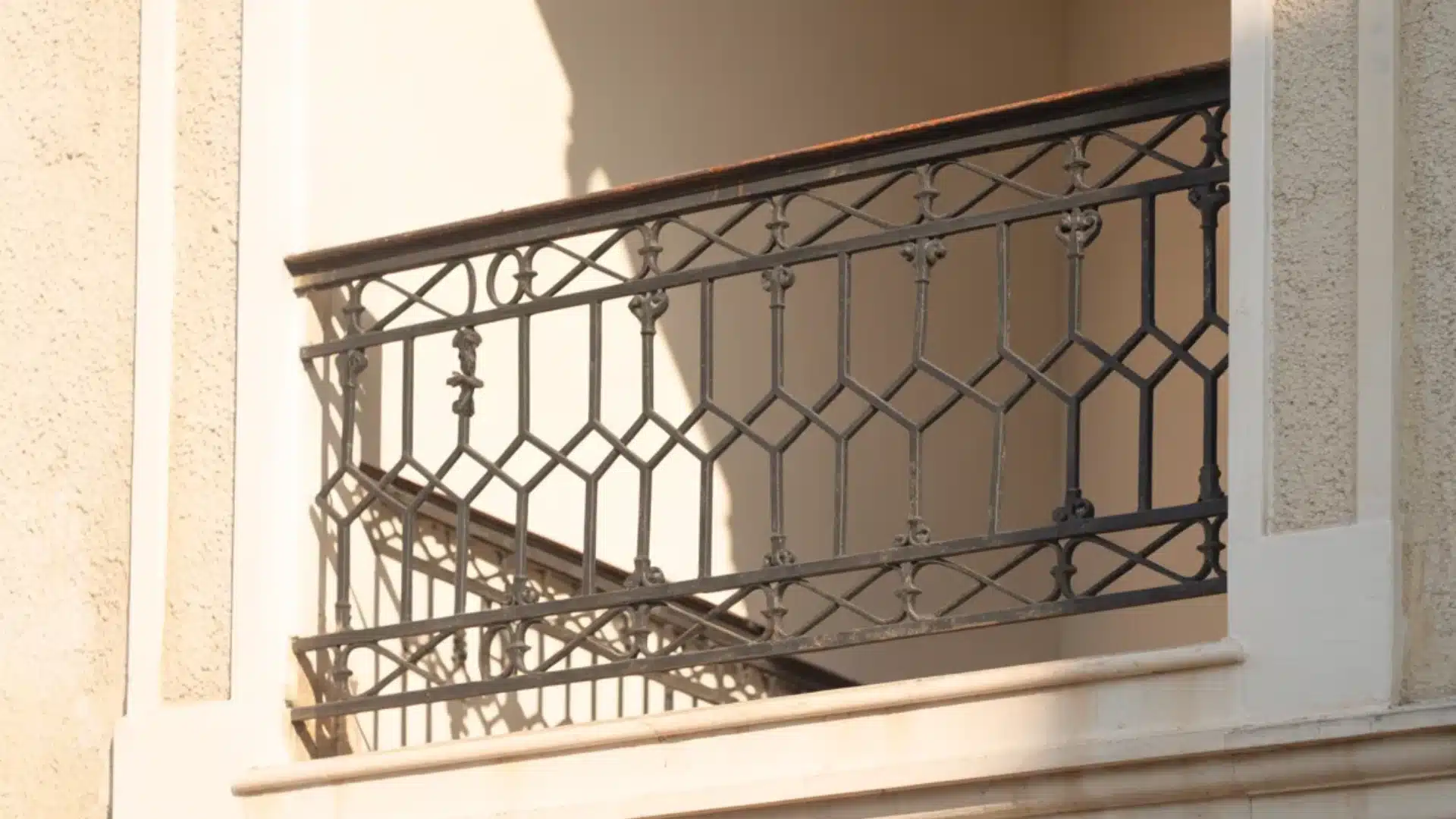 Close up of a black metal balcony railing with ornate geometric patterns on a beige building with bright sunlight cast over