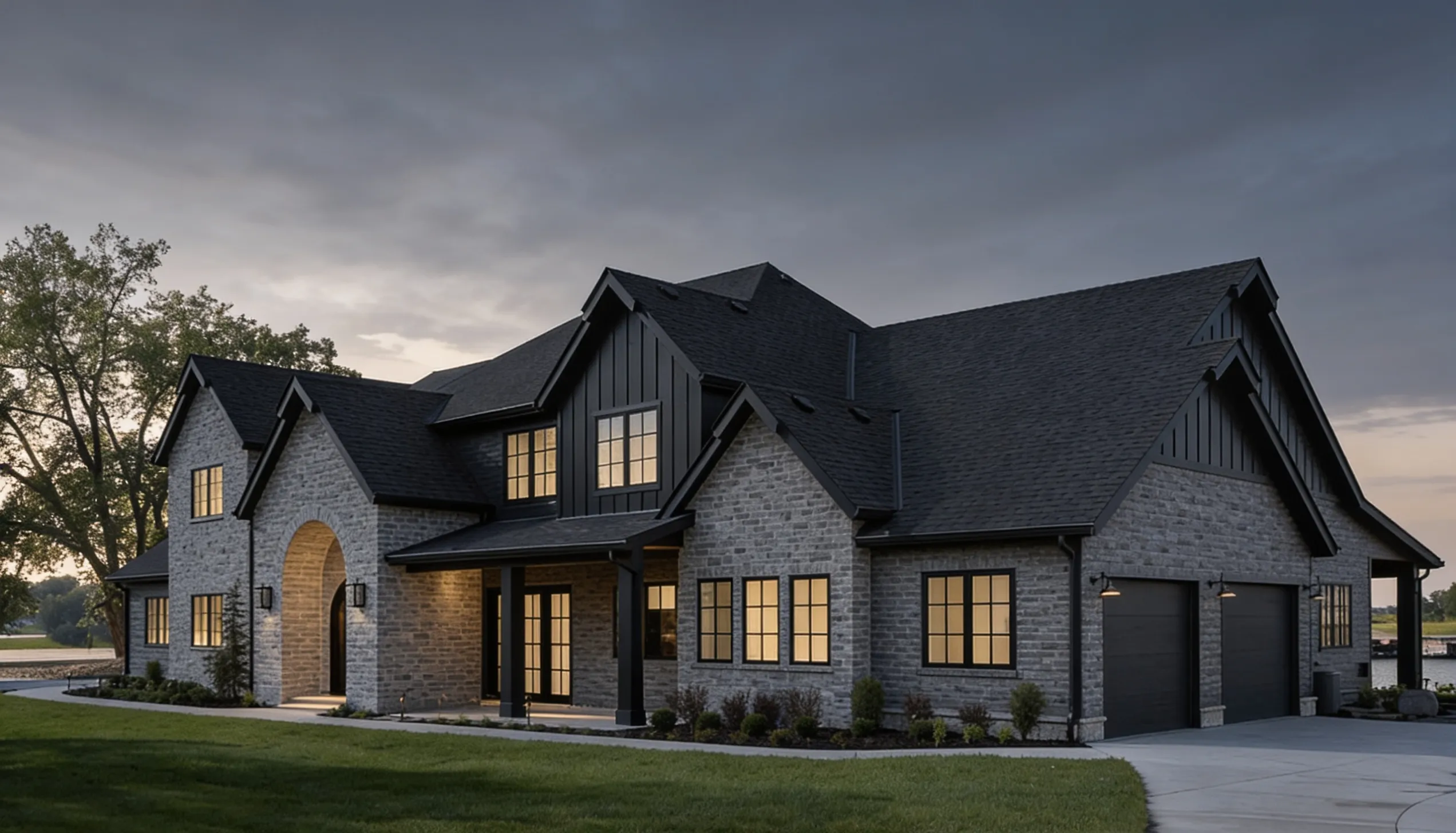 Large custom home featuring grey stone exterior, black roof, black trim, and warm interior lighting against a dark, overcast sky.