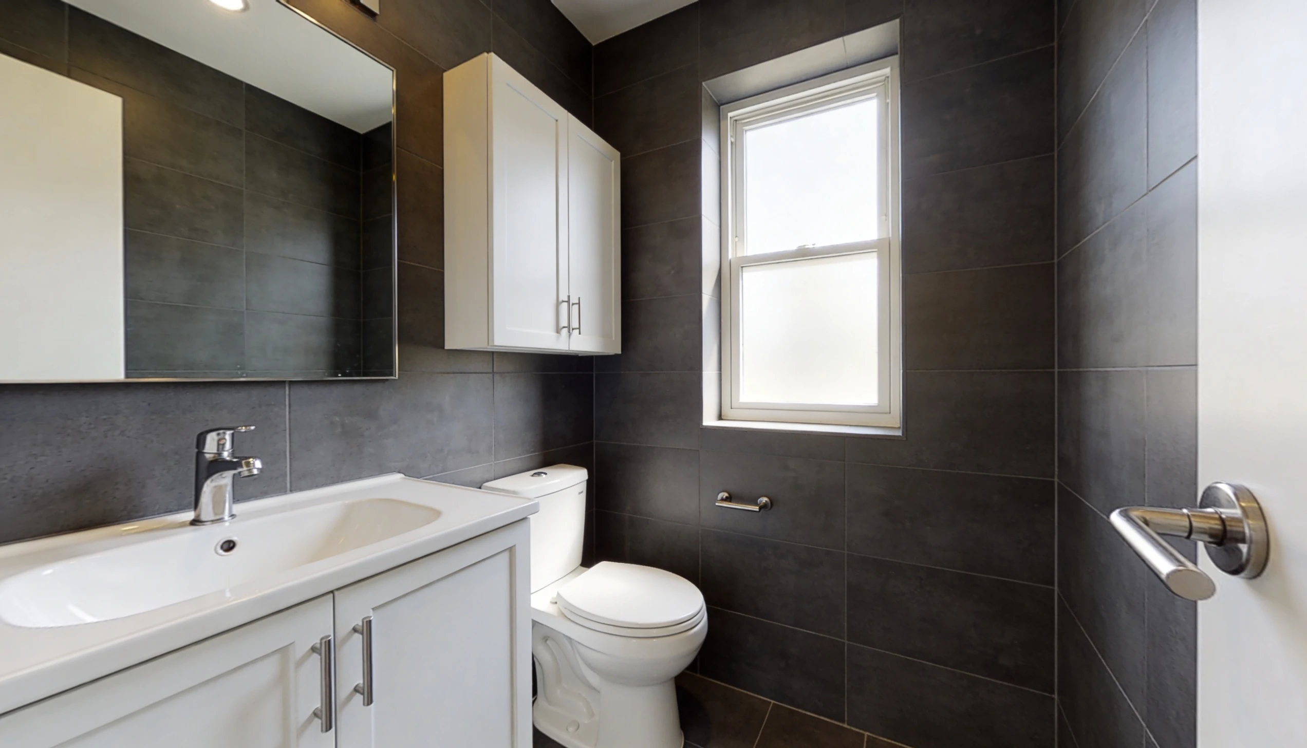 Modern bathroom with charcoal grey tiled walls, white vanity and toilet, bright window light, showing clean interiors